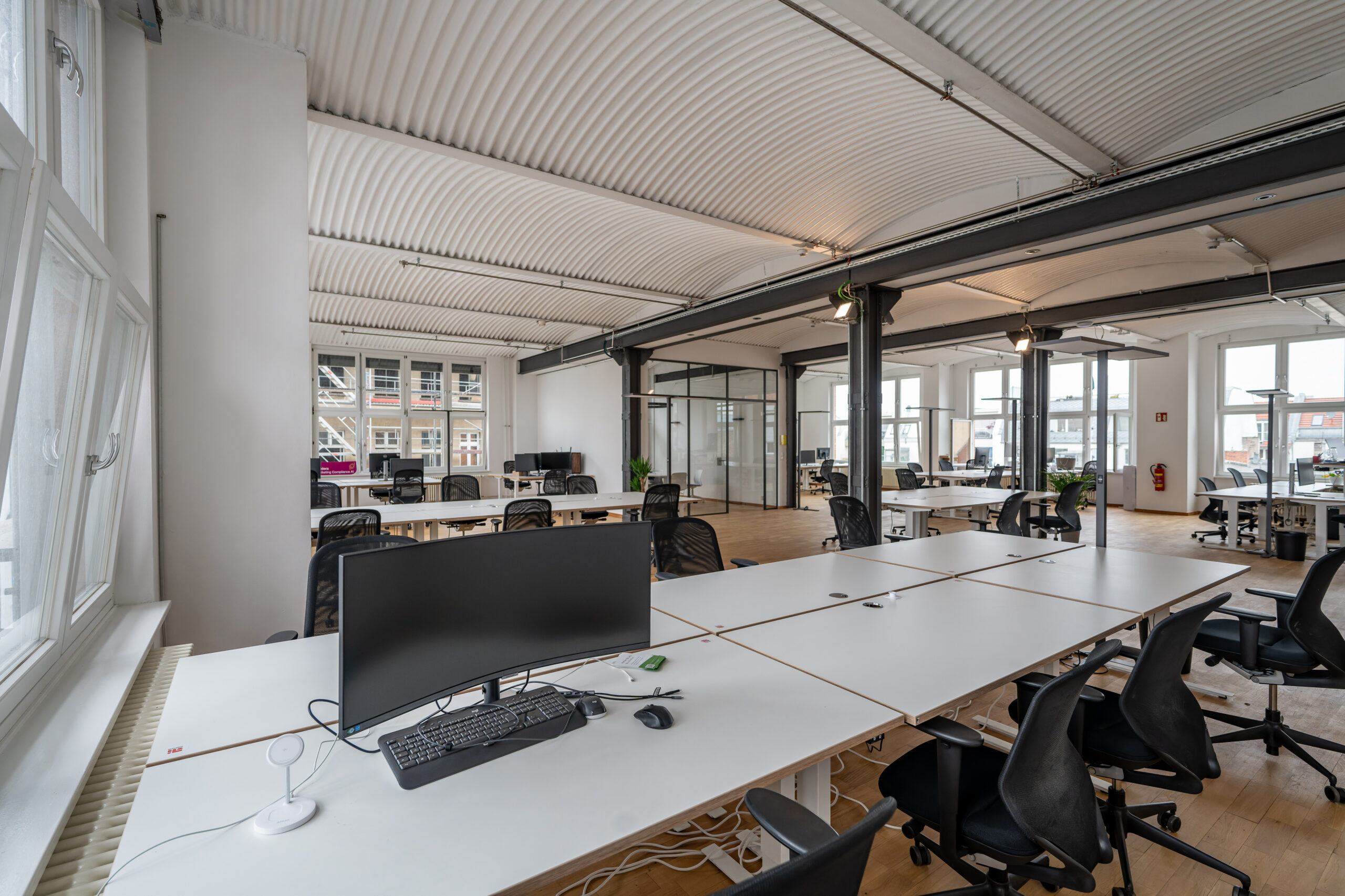 Modern open-plan office with white desks and corrugated ceiling.